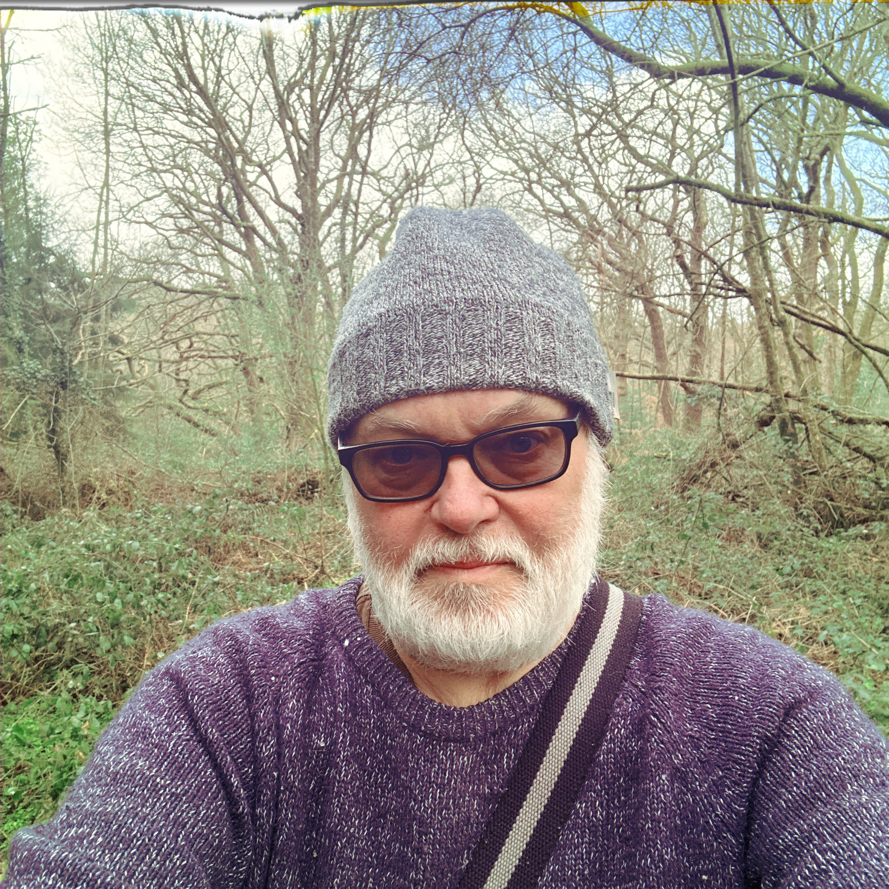 Auto-generated description: A person wearing a gray beanie and sunglasses takes a selfie in a wooded area.