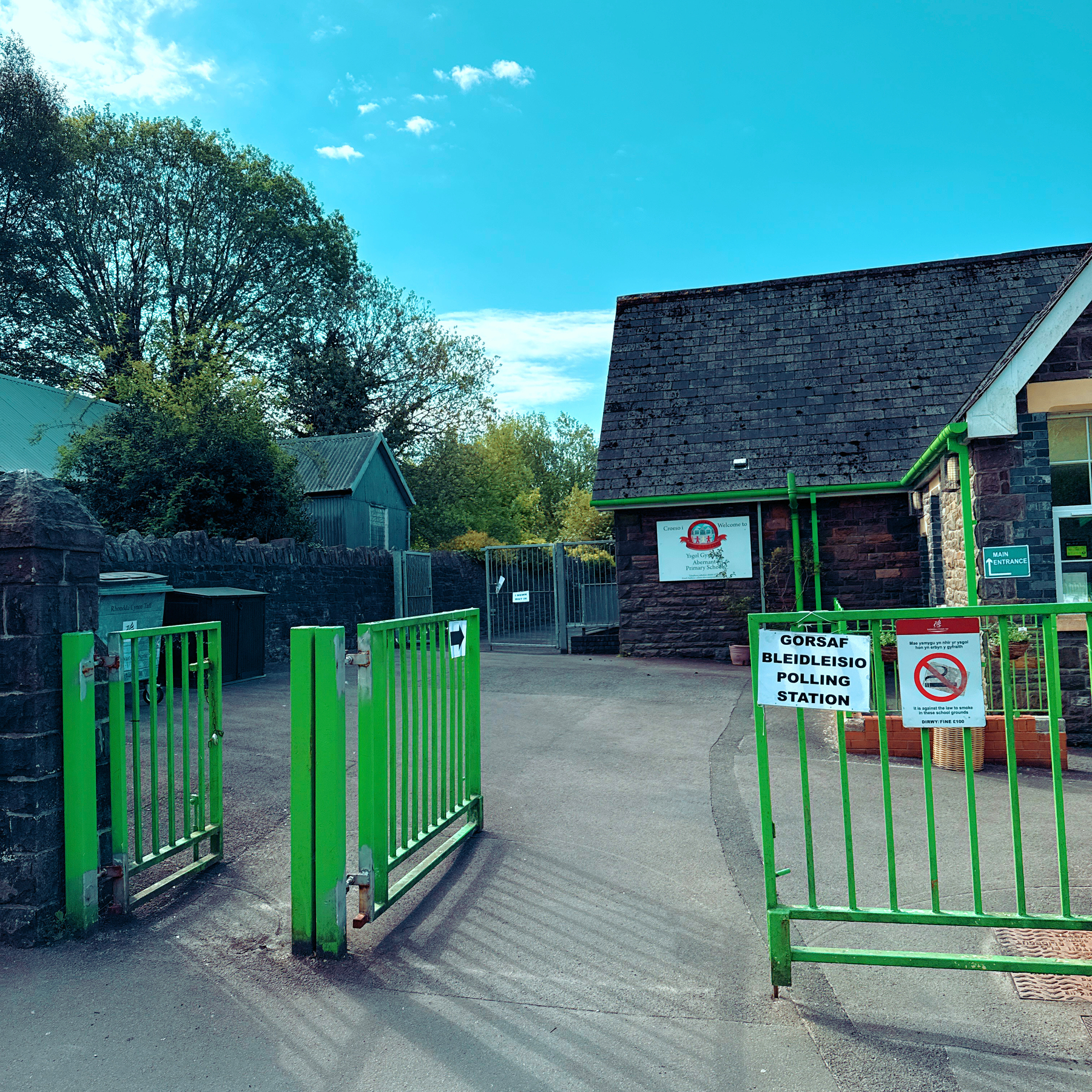 A small brick building with signs indicating it's a polling station is situated behind green gates on a clear day.