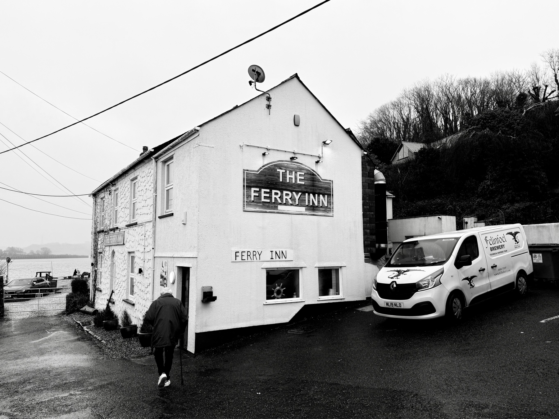 Auto-generated description: A person walking by a waterfront building labeled The Ferry Inn, with a parked van nearby on an overcast day.