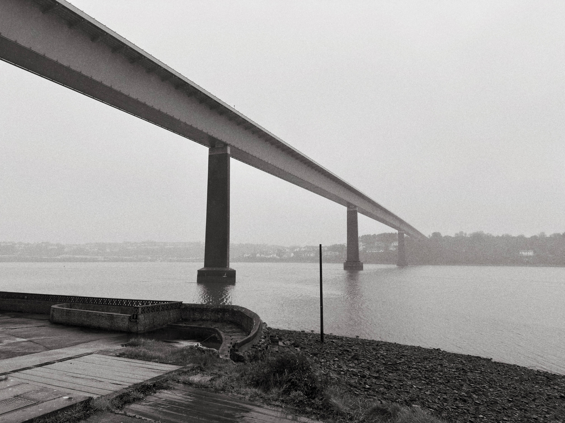 Auto-generated description: A long bridge stretches across a foggy body of water, supported by tall pillars, with a shoreline in the foreground.