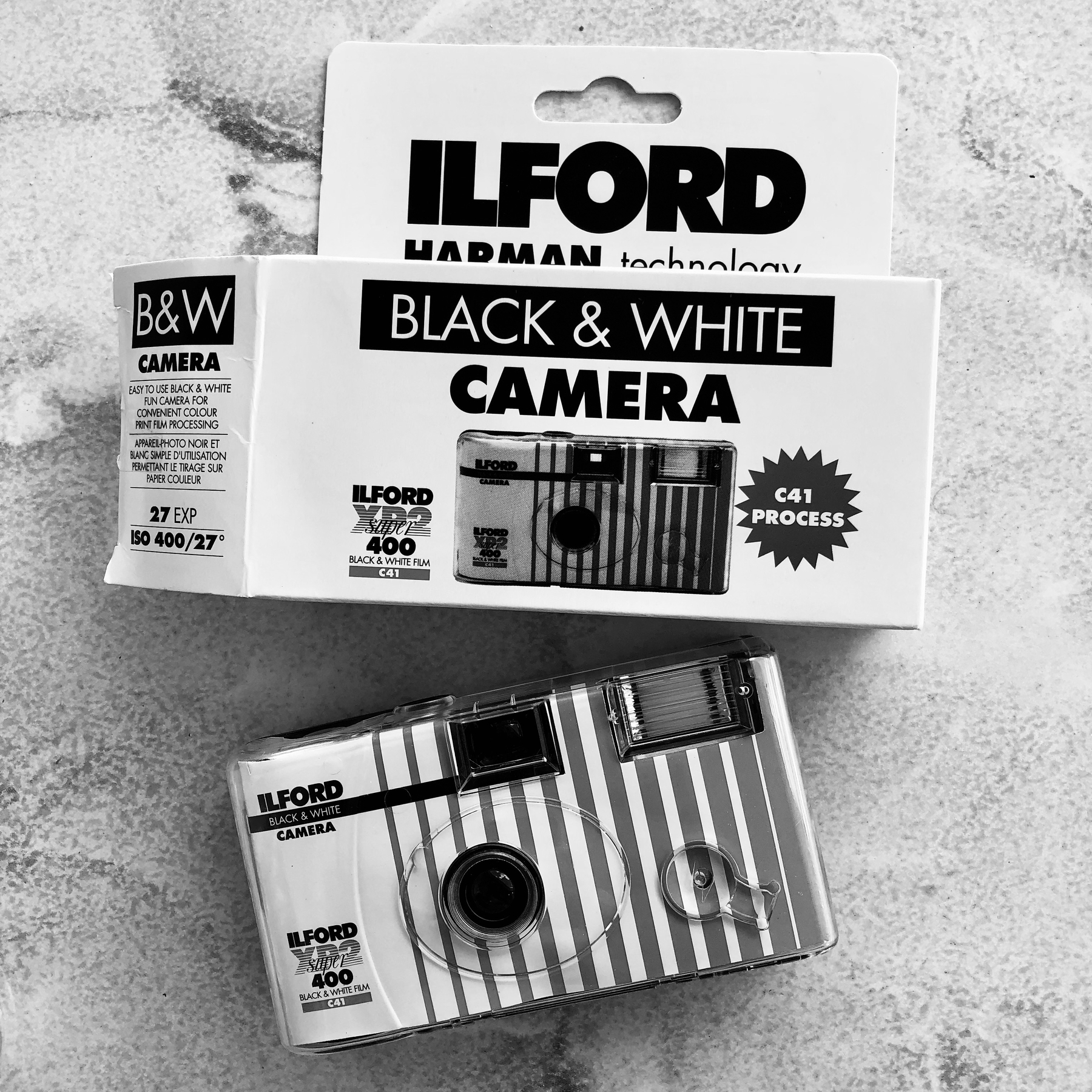 A black and white disposable camera with its box labeled Ilford Black & White Camera is displayed on a textured surface