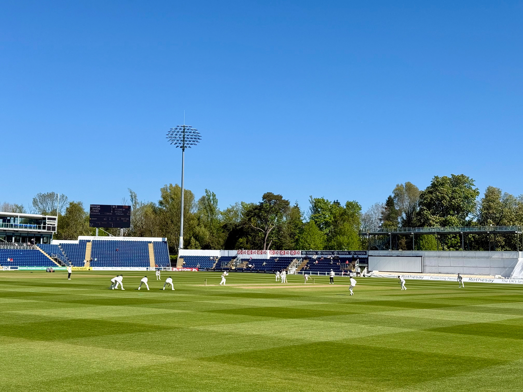 A cricket match is taking place on a sunny day in a stadium surrounded by trees.