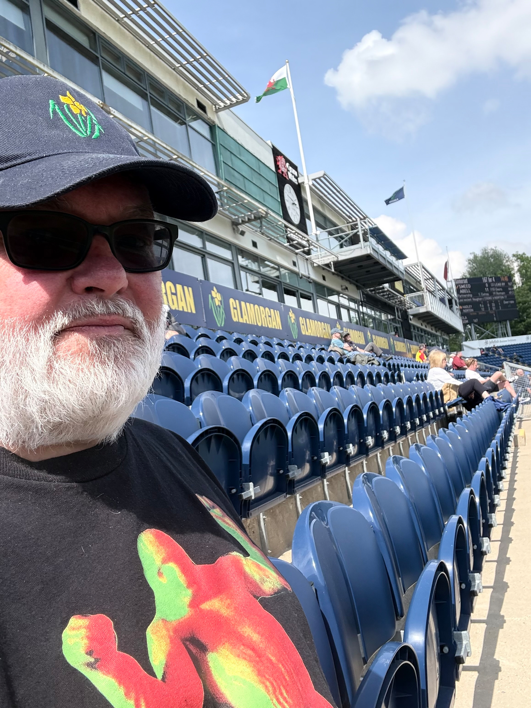 A bearded man wearing a cap and sunglasses sits in a stadium with rows of blue seats under a partly cloudy sky.