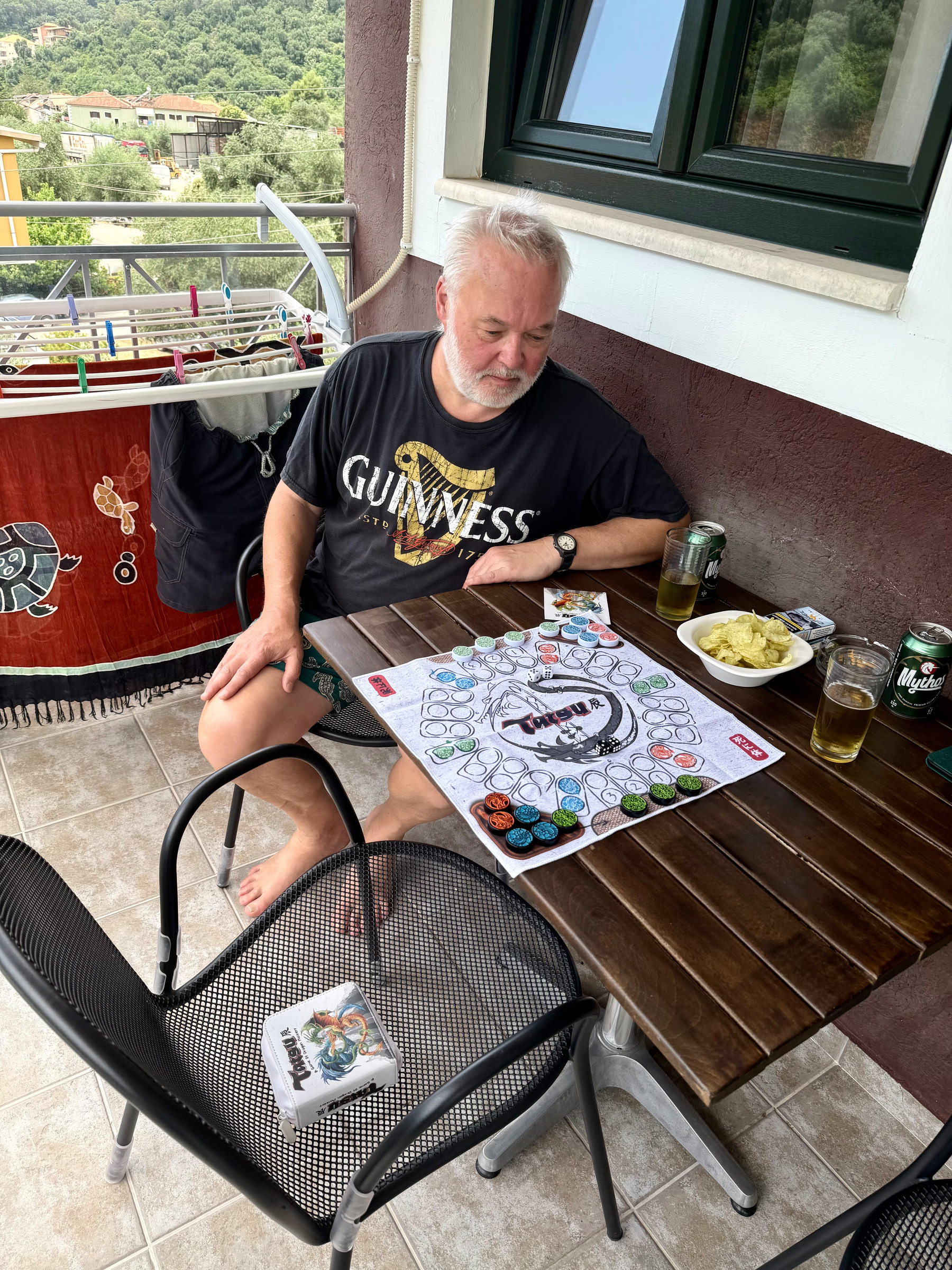 A man wearing a Guinness t-shirt is sitting at a table on a balcony playing a board game named Tatsu, with snacks and drinks beside him.