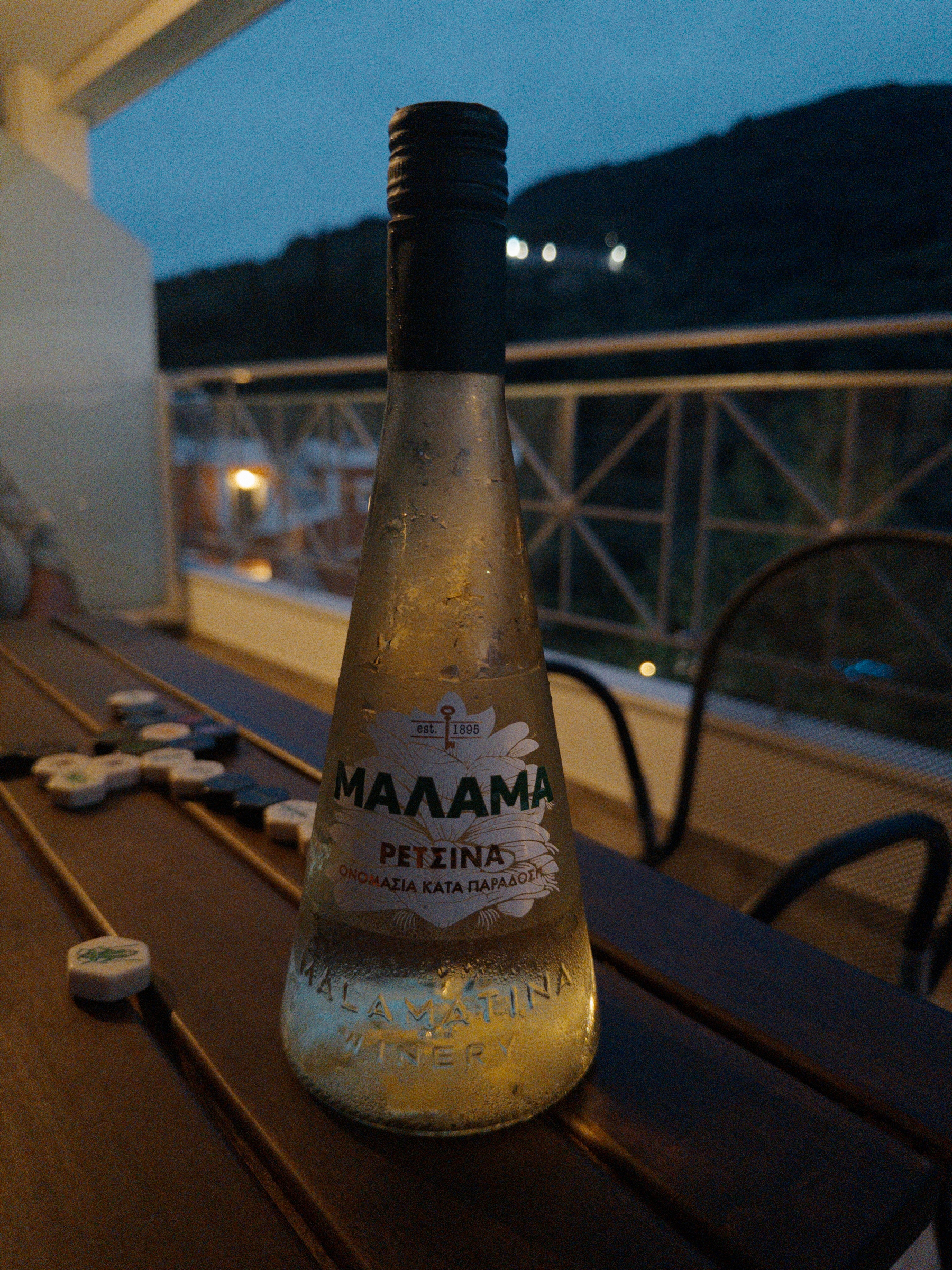 A frosted bottle of Malamatina Retsina wine is placed on a wooden table outdoors, with a scenic evening background.