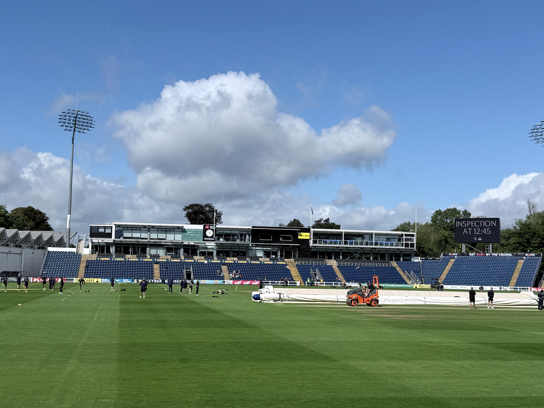 A cricket ground with players on the field, a covered pitch, and a scoreboard reading Inspection at 12:45 under a partly cloudy sky.