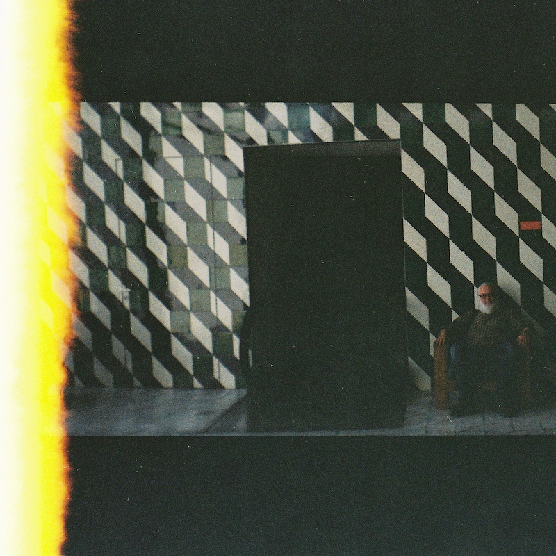 profile image - a bearded man is sitting on a chair against a geometric patterned wall with a dark passageway beside him and a light leak effect on the left side of the photo